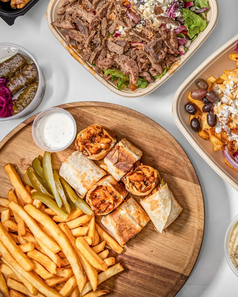 Chicken shawarma bites and fries on a wooden board next to a beef plate, grape leaves and loaded fries