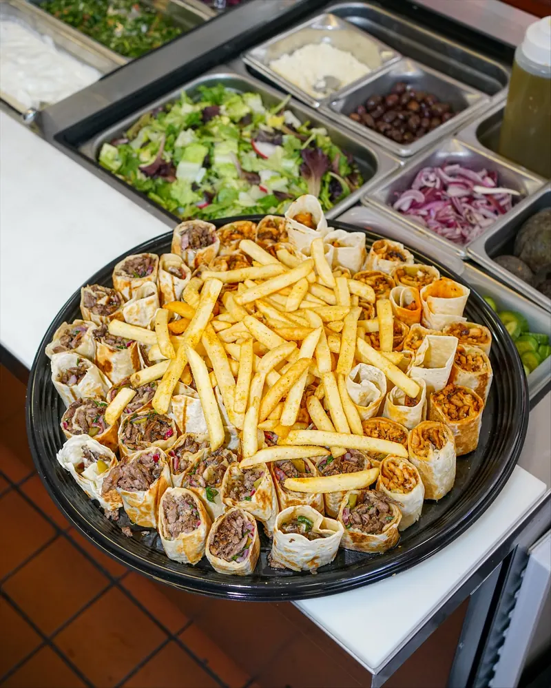 A catering tray of shawarma bites topped with fries, with salad trays behind