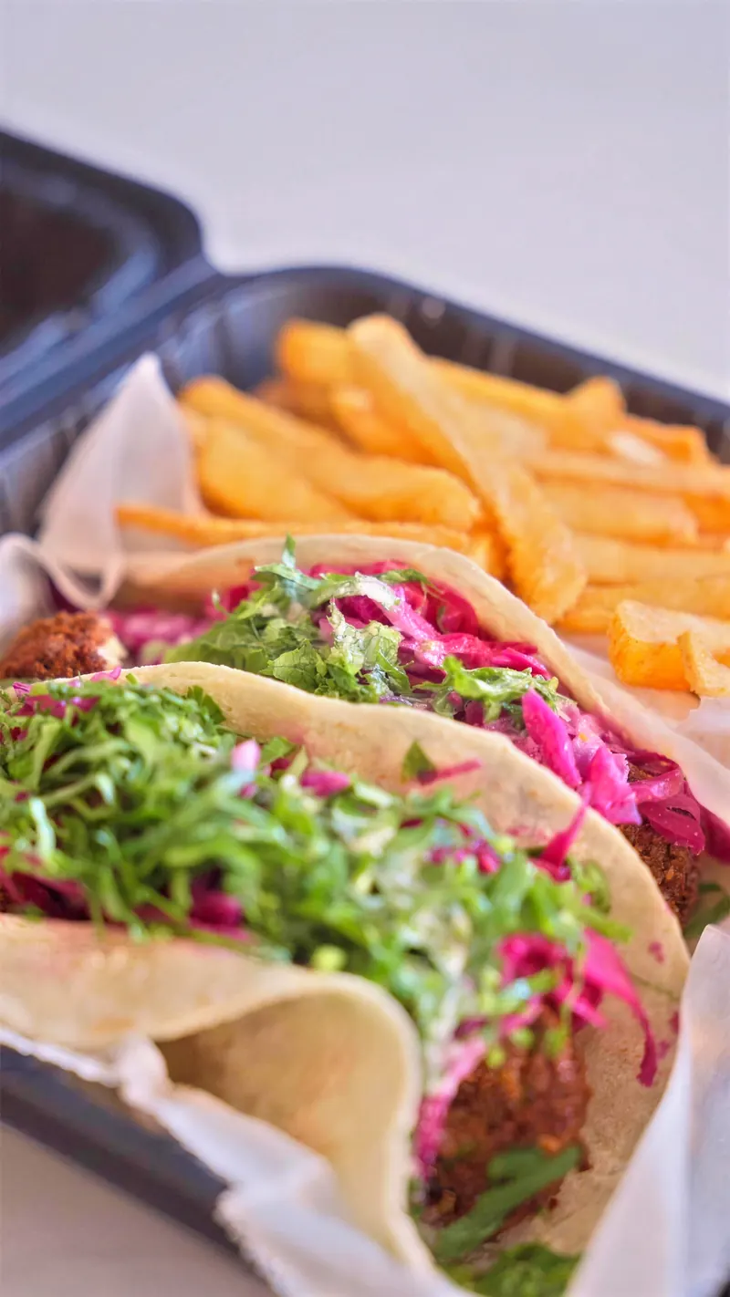 Two spicy falafel tacos with greens and pickled cabbage, fries behind