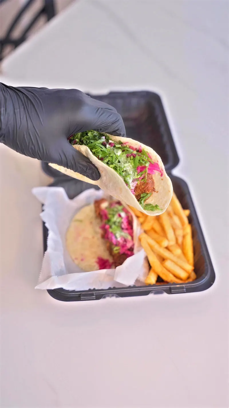A spicy falafel taco held over an open to-go box of fries