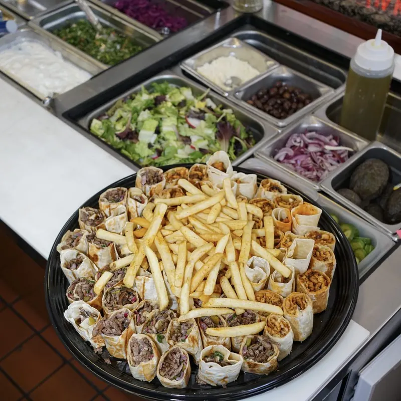 A catering tray of shawarma bites topped with fries, with trays of salad behind