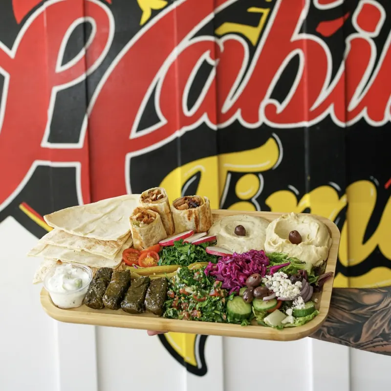 A starters board with hummus, shawarma bites, grape leaves, tabbouleh and pita, held up in front of the mural